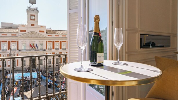Een balkon met uitzicht op een historisch gebouw, een champagnefles en twee glazen op een tafel.