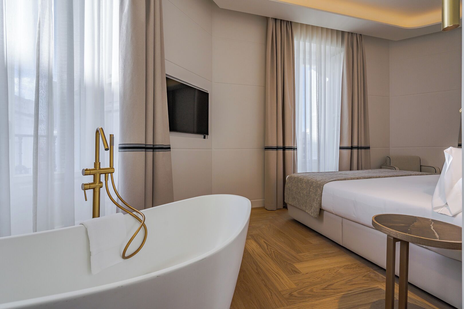 A modern hotel room with a bathtub, a bed, a TV mounted on the wall, and a small table.