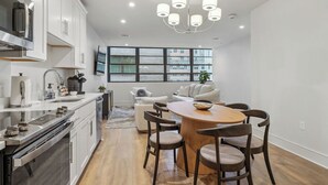 Una cocina moderna con electrodomésticos de acero inoxidable, una zona de comedor con una mesa y sillas de madera, y un ventanal con vistas a un edificio.