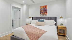 Un dormitorio con cama, ventilador de techo, un cuadro, lámparas de noche y una planta pequeña.