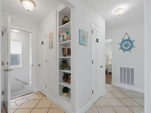 A hallway with white walls, a tiled floor, and a door leading to another room. There are shelves with various items and a decorative wheel on the wall.