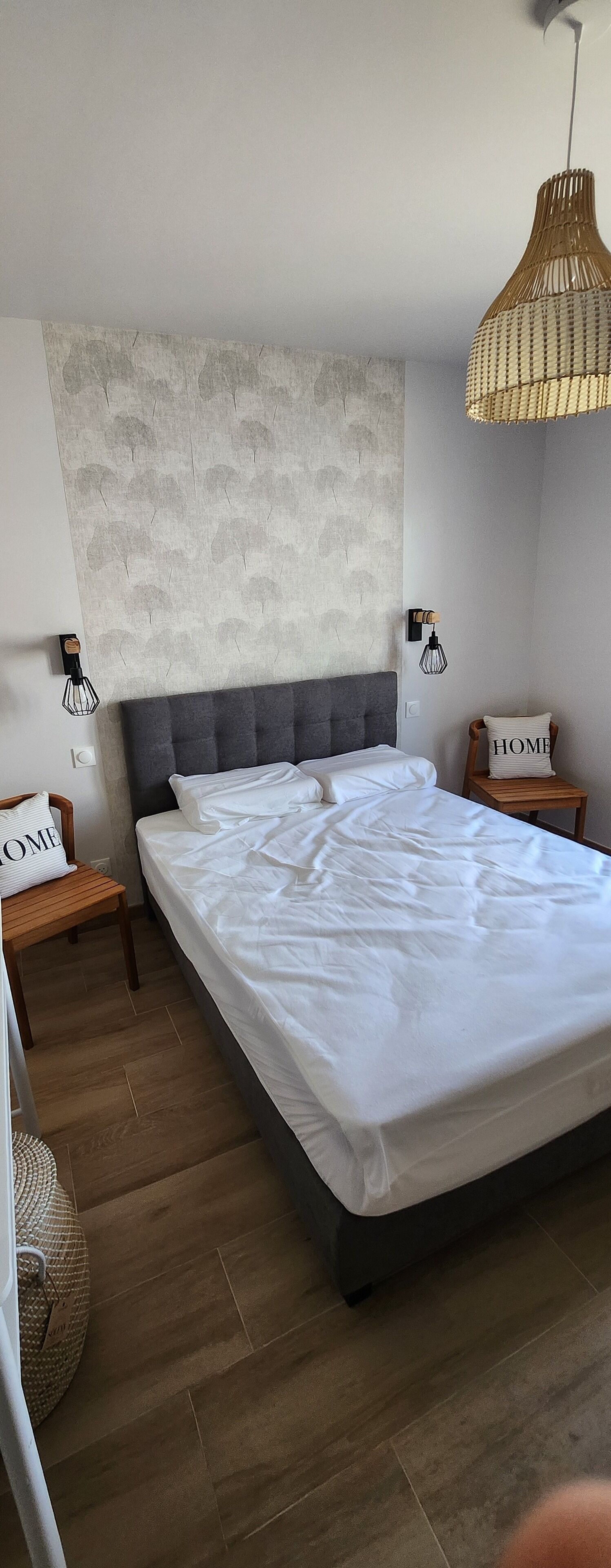 Ein Schlafzimmer mit einem großen Bett, einem grau gepolsterten Kopfteil, einer strukturierten Wand, einer gewebten Pendelleuchte und zwei Nachttischen mit Kissen mit der Aufschrift 'HOME'.
