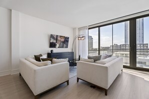 A modern living room with two white sofas, a dark-colored cabinet, and a large window offering a city view. - The Theodore 5BD/5B - Combo Unit - 605/606 (New Orleans)