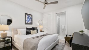 Ein modernes Schlafzimmer mit Bett, Nachttischen, Deckenventilator, einem gerahmten Kunstwerk und einer Tür zu einem weiteren Raum.