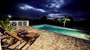 A swimming pool with stone flooring, a wooden lounge chair, and a white fence.