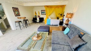 Un salon chaleureux avec un canapé, une table basse et un lit surmonté d’un dais jaune. On peut voir des planches de surf accrochées au mur et une affiche d’Eddie Aikau.