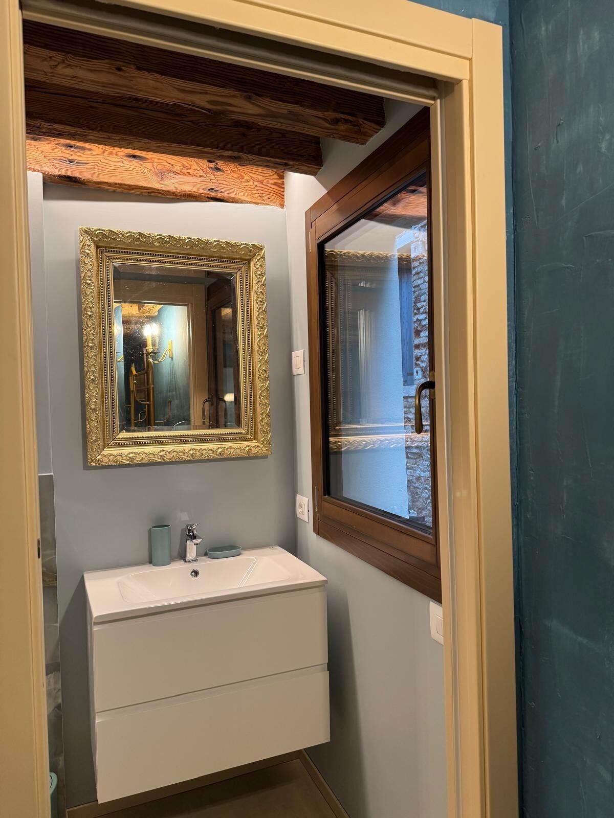 Ein Badezimmer mit einem weißen Waschbecken, einem spiegel mit goldfarbener Rahmung und einem Fenster mit Blick auf ein Backsteingebäude.