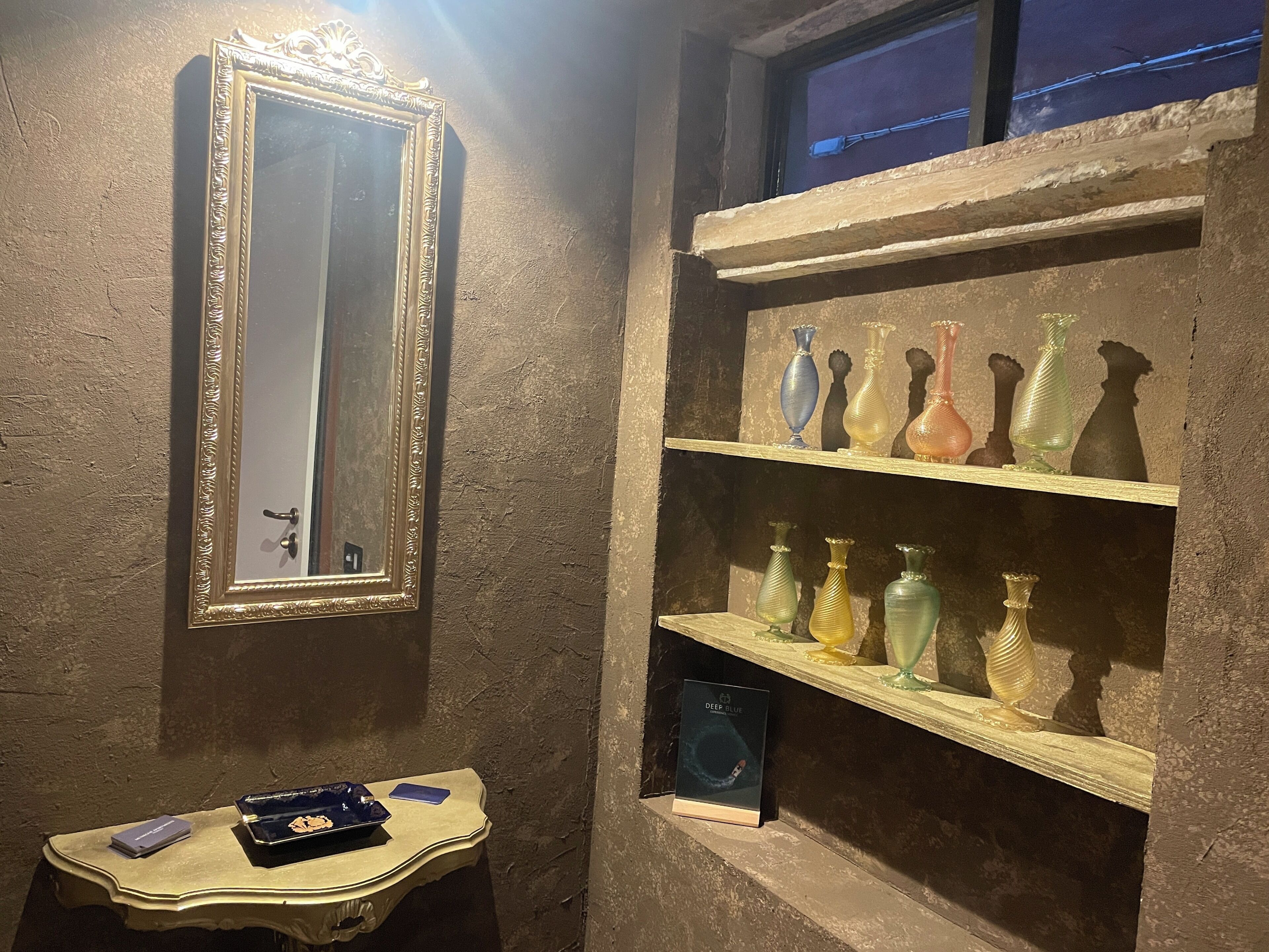 Un baño con espejo, estantes con jarrones, una tocador y una ventana.