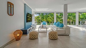 Un salon moderne avec un téléviseur, des canapés blancs, des poufs à motifs et une grande fenêtre donnant sur un jardin.