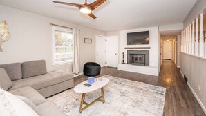 A living room with a sofa, a coffee table, a ceiling fan, a fireplace, and a hallway with a railing. - The Terrace Timber House  For 8 (Hamilton)