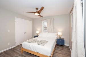 Un dormitorio con cama, ventilador de techo, ventana con cortinas, mesita de noche con lámpara y puerta.