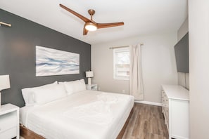 A bedroom with a bed, nightstands, a ceiling fan, a window with curtains, and a painting on the wall.