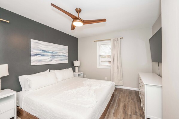 A bedroom with a bed, nightstands, a ceiling fan, a window with curtains, and a painting on the wall.