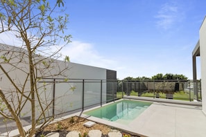A modern outdoor pool area with a glass enclosure, a tree, and a view of a residential building in the background.