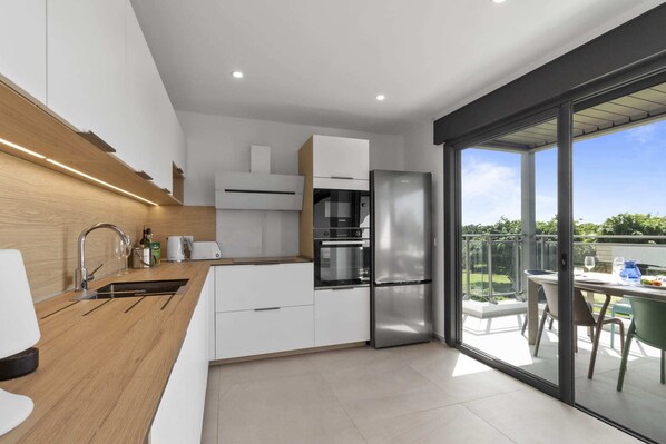 Una cocina moderna con armarios blancos, electrodomésticos de acero inoxidable y un amplio ventanal corredero que da a un patio exterior.