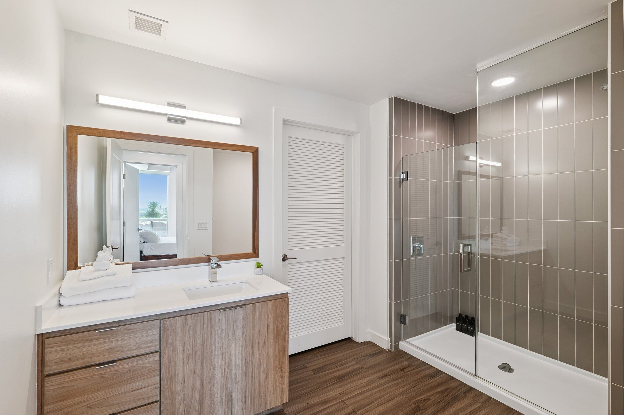 A modern bathroom with a wooden vanity, a large mirror, a glass-enclosed shower, and a white door.