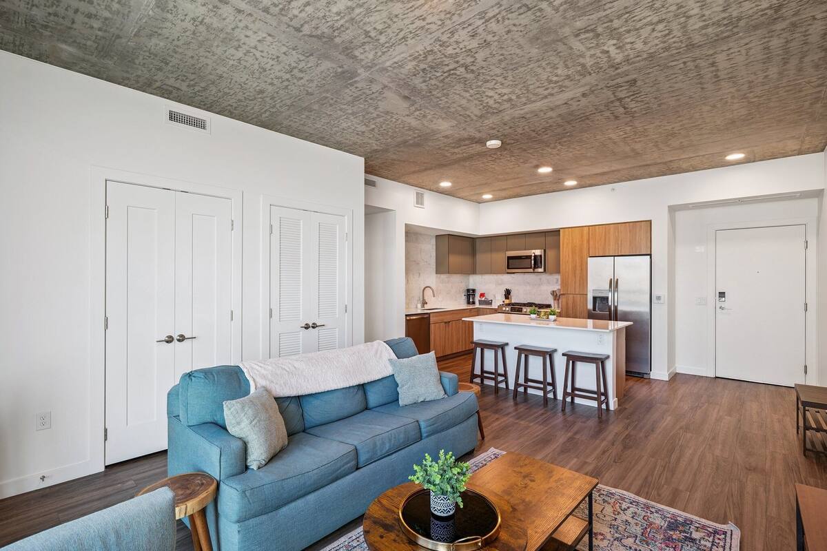 Una sala de estar moderna con un sofá azul, una mesa de centro de madera y una cocina con una isla central, taburetes de bar y electrodomésticos integrados.
