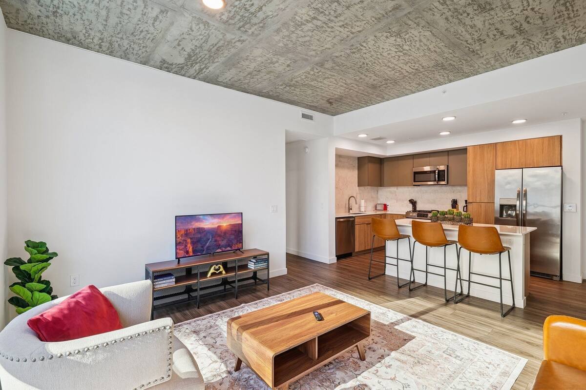 Una sala de estar moderna con sofá, mesa de centro y televisor, conectada a una cocina con taburetes y electrodomésticos.