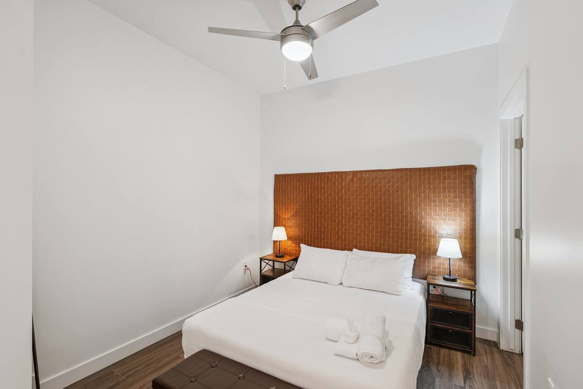 Habitación de hotel con cama, dos mesitas de noche con lámparas, ventilador de techo y cabecero de madera.