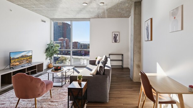 A modern living room with a grey sofa, a wooden dining table, and a television on a stand. - CozySuites at ADELINE (Phoenix)