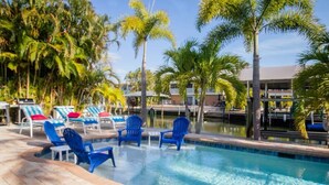 Un área de piscina con sillas y sillones azules, rodeada de palmeras y un edificio al fondo.