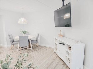 Ein Essbereich mit rundem Tisch, grauen Stühlen, einer hängenden Leuchte, einem weißen Konsolentisch und einem an der Wand befestigten Fernseher.