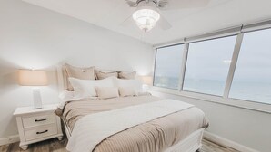 Une chambre à coucher comprenant un lit, des tables de chevet, un ventilateur de plafond et une grande fenêtre donnant sur un beau paysage.