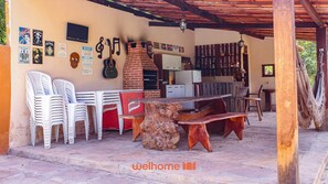 Un espace extérieur abrité comprenant un four en briques, des chaises en plastique blanches, un réfrigérateur et une table en bois agrémentée d’un centre de table fait d’une souche d’arbre.