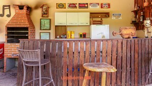 A rustic outdoor bar area with a brick oven, wooden furniture, and various beer and liquor bottles displayed on shelves. - Suite Quadrupla em Rio Grande com Vista Montanha (Palmeiras)