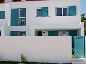 En moderne bygning med to etager, teal farvede skodder på vinduer og døre, et fladt tag og en hvid facade.