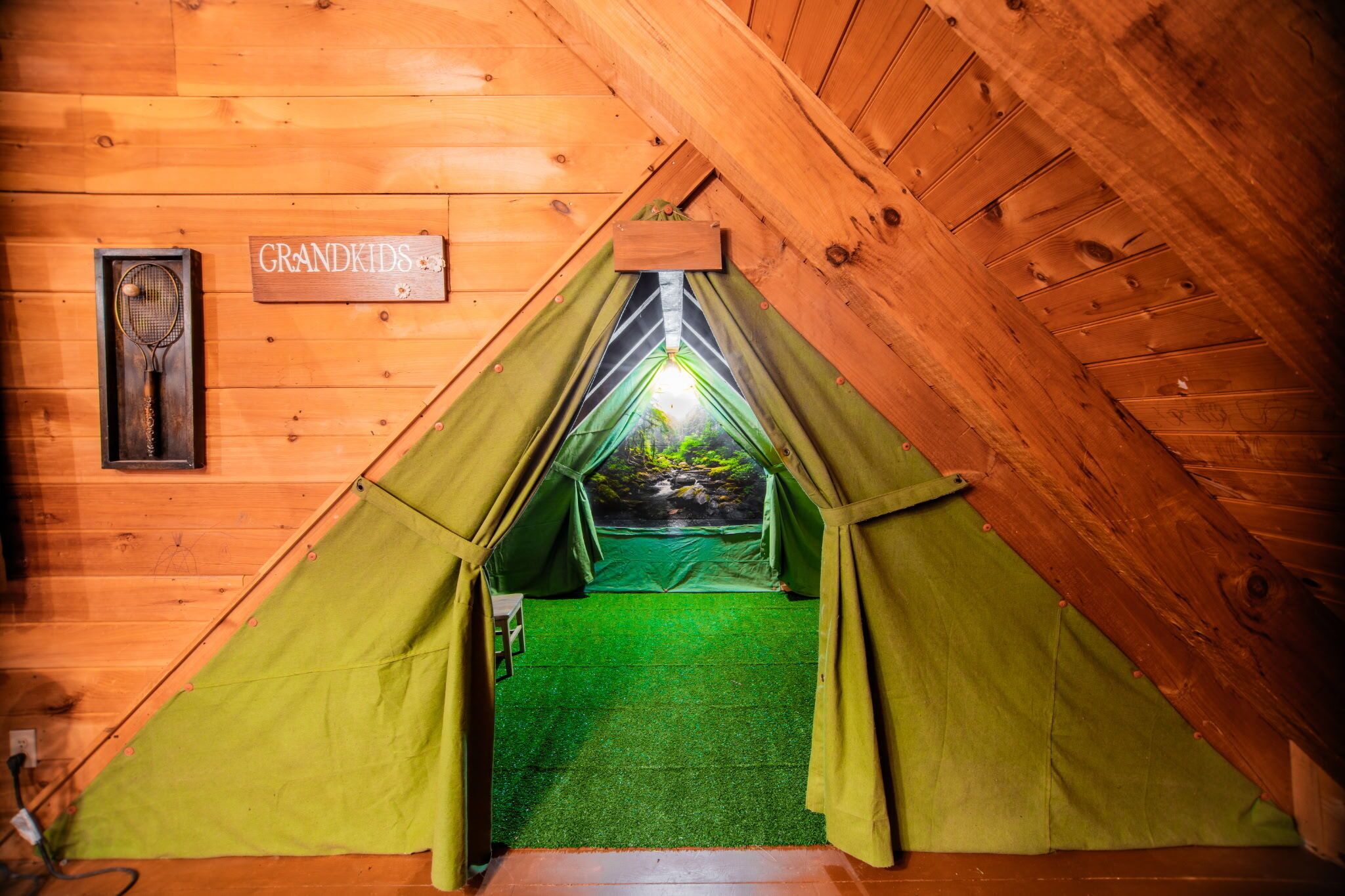 Una cabaña de madera con entrada tipo tienda de campaña verde, un letrero de madera que dice "GRANDKIDS" y una raqueta de tenis colgada en la pared.