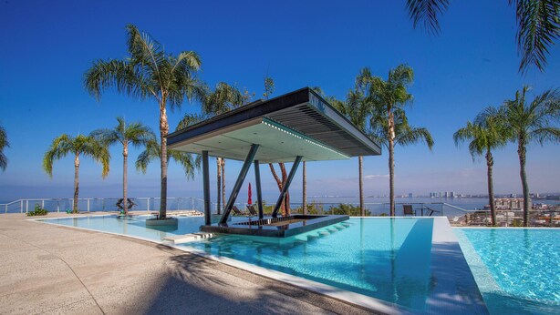 Ein moderner Infinity-Pool mit überdachter Sitzgruppe, umgeben von Palmen und einer Stadtkulisse im Hintergrund.
