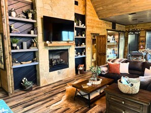 Ein gemütliches Wohnzimmer mit Kamin aus Stein, Holzregalen, einem Fernseher und einer bequemen Sitzecke.