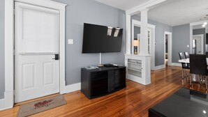 Una sala de estar moderna con un televisor de pantalla plana, piso de madera, mueble de entretenimiento negro y un comedor con una mesa y sillas blancas.