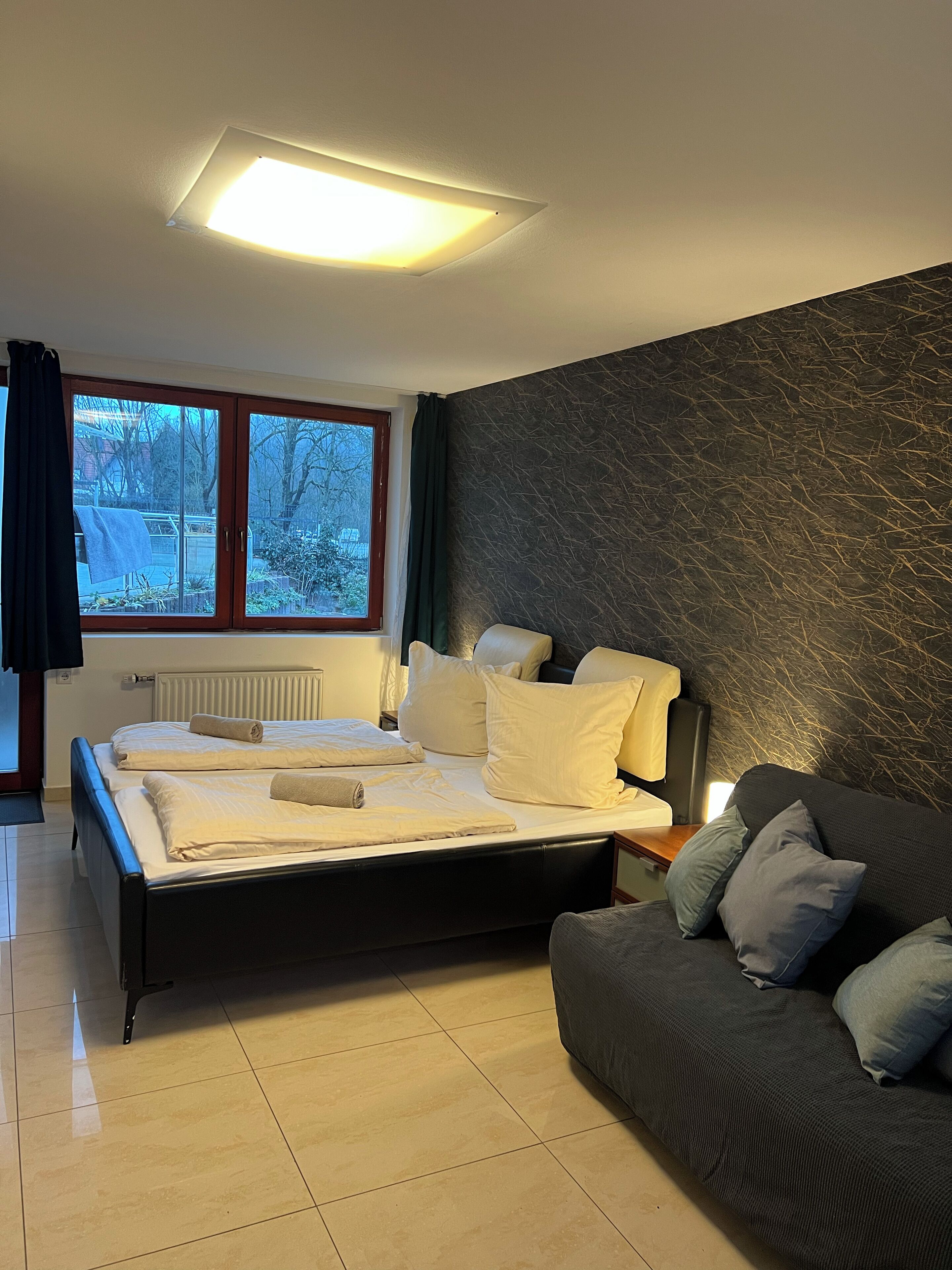 A bedroom with a bed, a dark-colored sofa, a window with curtains, and a radiator.