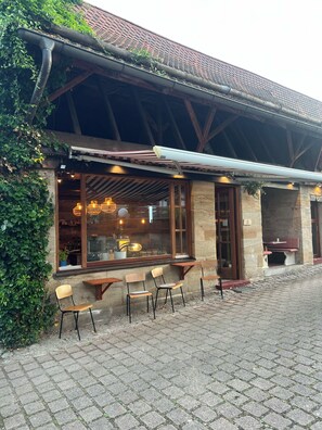 A rustic outdoor seating area with wooden tables and chairs, a stone wall, and a covered roof. - Hotel Scheunenwerk (Heroldsberg)