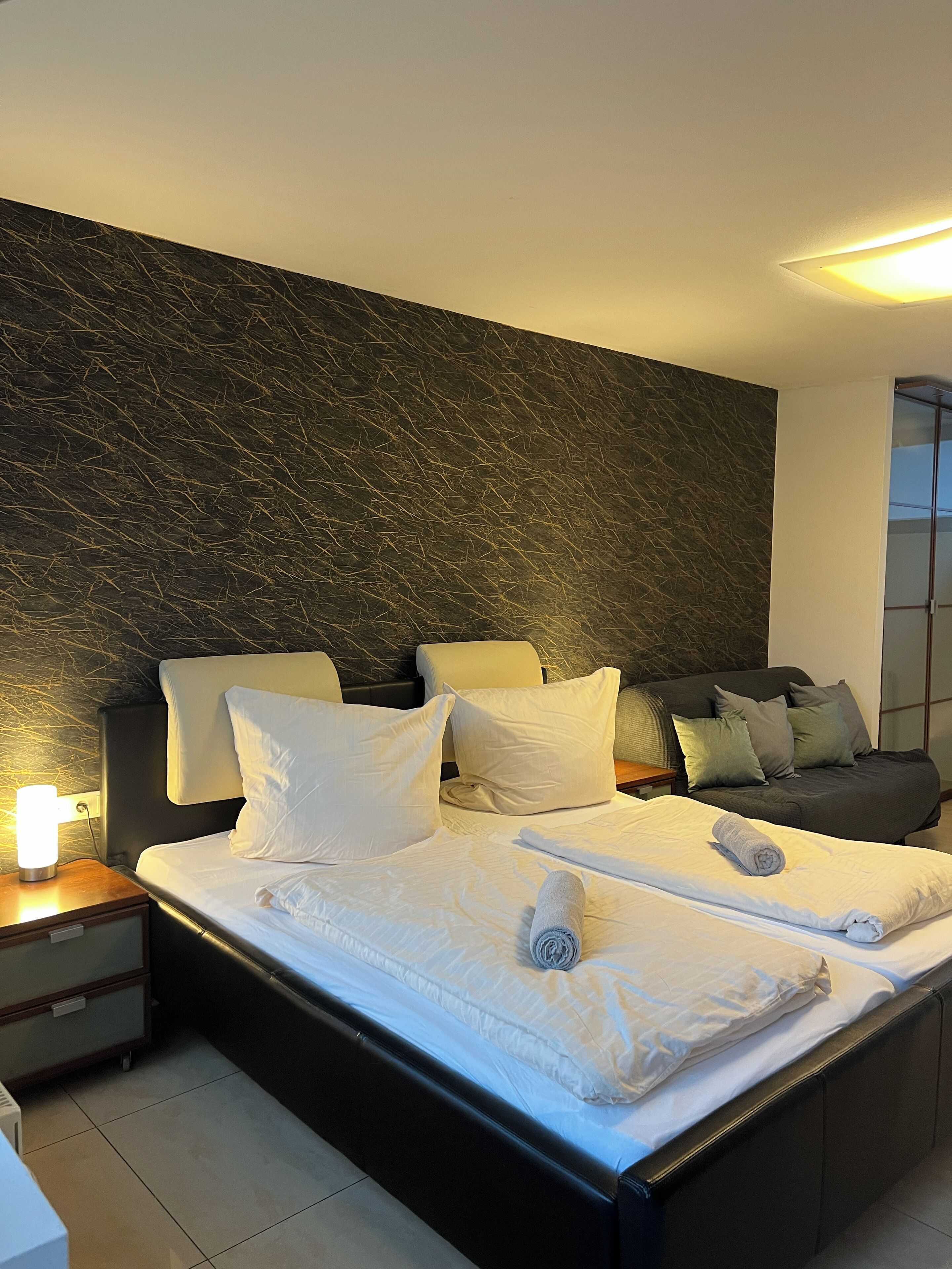 Una habitación de hotel moderna con una cama grande, una mesita de noche, un sofá y una pared con un diseño texturizado.