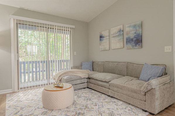 A living room with a beige sectional sofa, a round ottoman, and a large window with blinds. There are three abstract paintings on the wall.