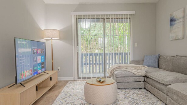 Una sala de estar con televisor, sofá, mesa de centro y vista a árboles a través de una ventana.