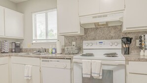 Una cocina con armarios blancos, estufa, lavavajillas y ventana con persianas.