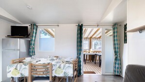 Ein Essbereich mit einem Tisch für vier Personen, Holzkästen, einem Fernseher auf einem Regal und einem durch eine offene Tür sichtbaren Küchenbereich.