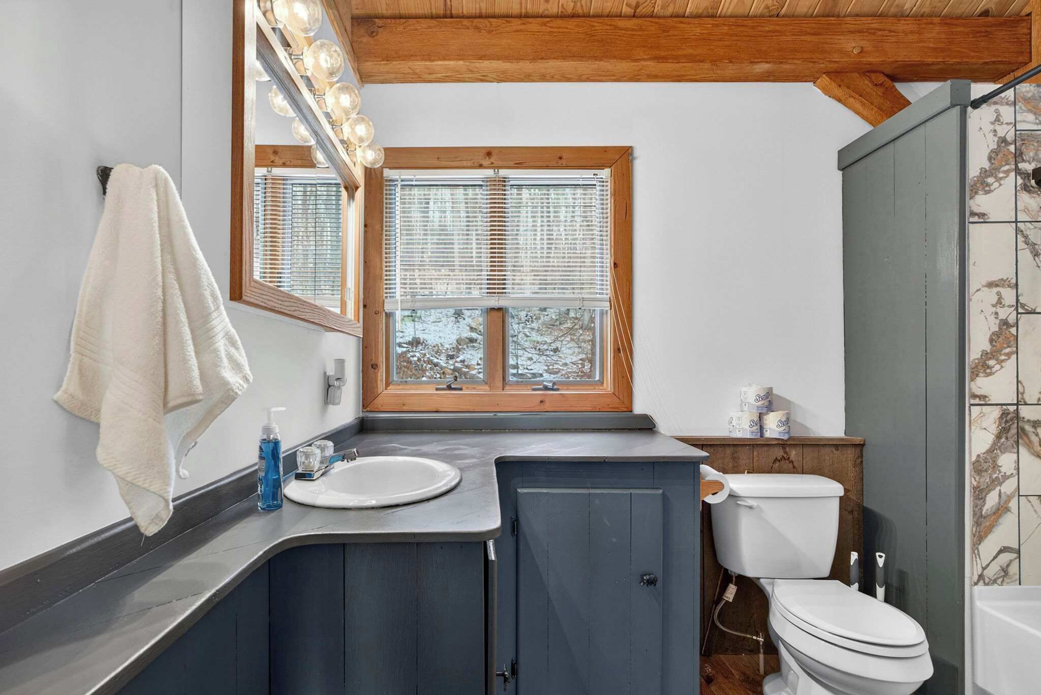 A bathroom with a vanity, sink, and toilet. There is a towel hanging on the wall and a window with blinds.