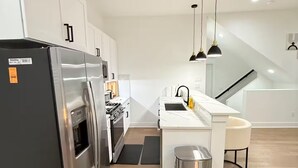A modern kitchen with a stainless steel refrigerator, white cabinetry, a built-in oven, and a stainless steel trash can. There are pendant lights above the kitchen island. - 3-bedroom house in serene Houston with cool AC for relaxing stays (Houston)
