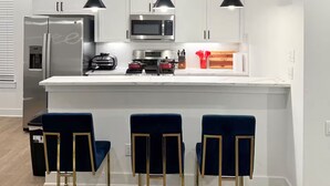 Una cocina moderna con armarios blancos, electrodomésticos de acero inoxidable y una isla central. Hay tres taburetes de bar azul oscuro con patas doradas.