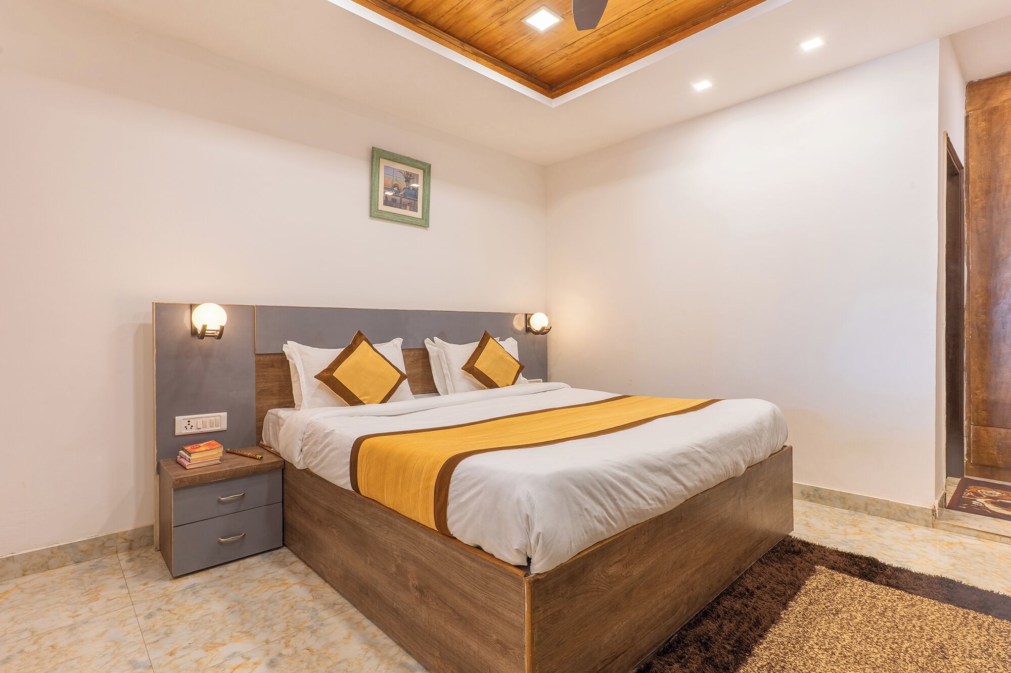 Een keurig opgemaakt bed met een houten hoofdbord, nachtkastjes met lampen en een ingelijst schilderij aan de muur.