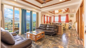 A spacious living area with a leather sofa, a glass coffee table, and a view of a mountainous landscape through large windows. - Lake Mount (Bhimtal)