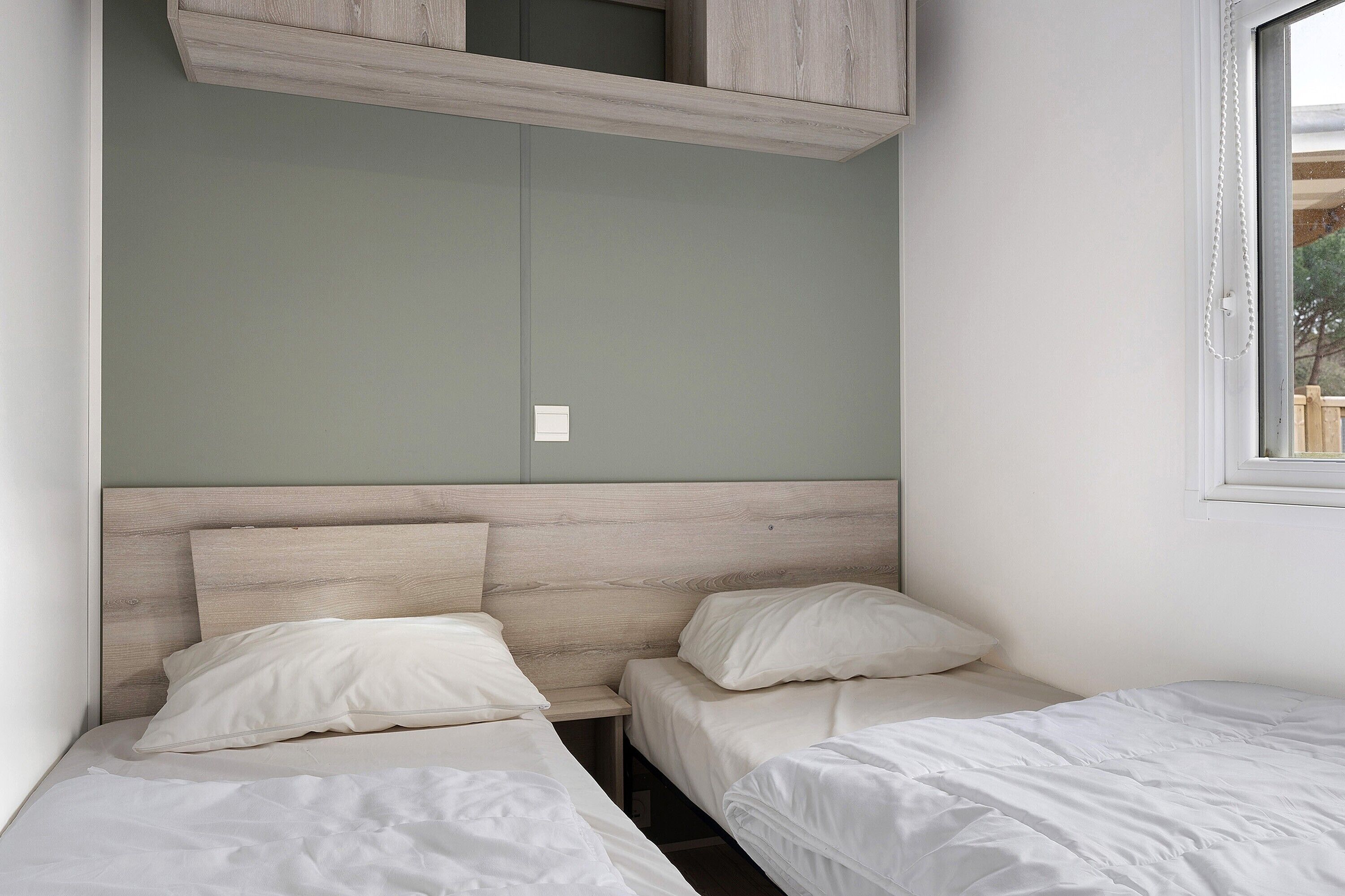 A room with two single beds, a wooden headboard, a small window, and a white wall.