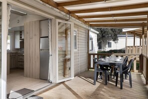 Um deck coberto com uma mesa posta para refeições, uma porta de vidro deslizante que dá para uma cozinha e um corrimão de deck em madeira.