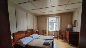 Una habitació amb dos llits, una finestra amb persianes, un radiador, un escriptori amb una làmpada i una cadira.