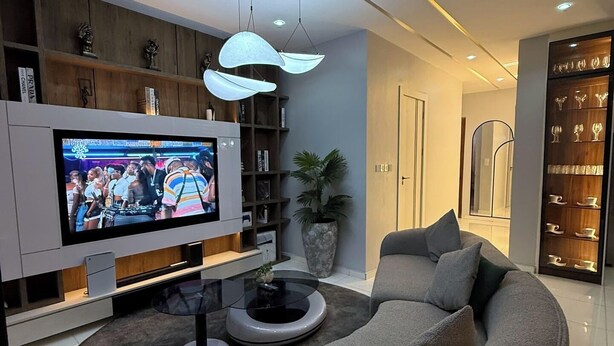 Una sala de estar moderna con un televisor de pantalla plana grande, un sofá gris curvo, una mesa de centro y una estantería.